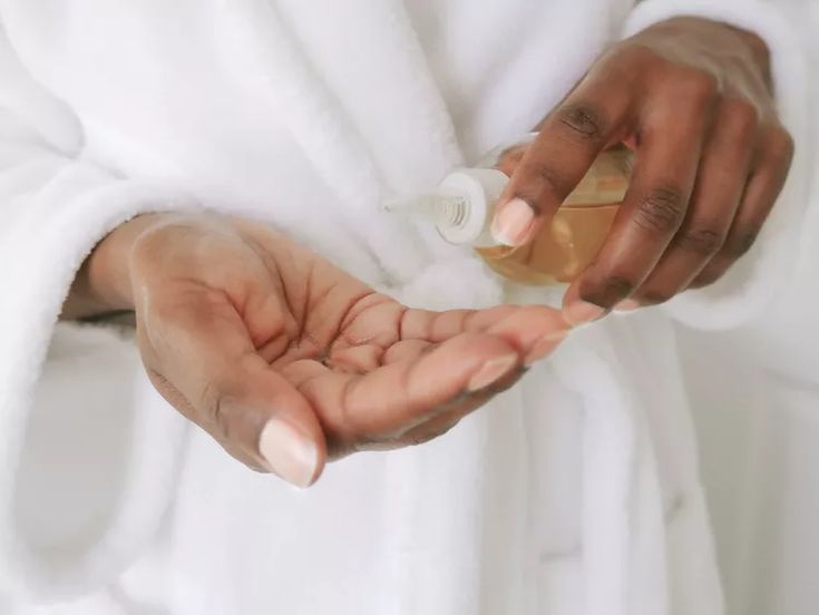 Black woman warming a few drops of hair oil between her palms before applying it evenly to damp sectioned 4C hair.