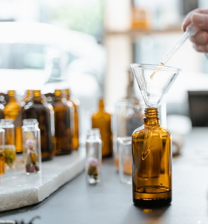 Several carrier oils in small glass bottles beside herbs, a dropper, and a mixing bowl.