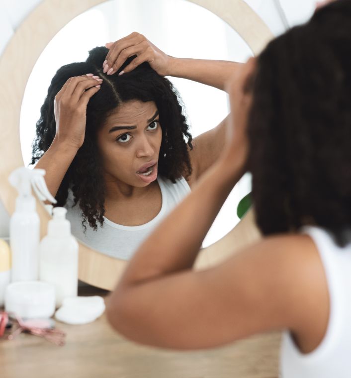 A woman with natural hair gently examining her scalp in a mirror.